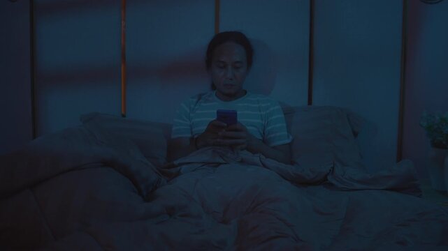 A man sits on a bed using a smartphone with the lights off at night, illustrating late-night screen time, bedtime scrolling, insomnia, and excessive social media use.