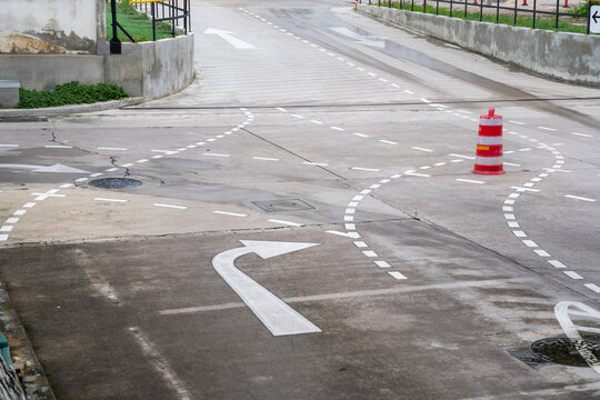 White directional arrows and dashed lane markings painted on a concrete road surface. Features a red traffic barrel and manhole covers in a driver training or testing area. Professional view.
