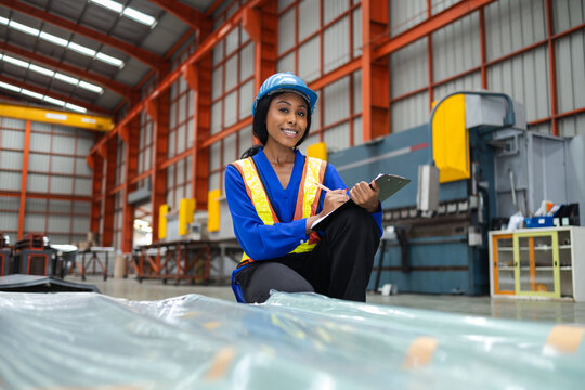 Happy smiling female factory worker wearing blue hard hat and yellow safety vest holding clipboard inspecting manufacturing warehouse industrial plant looking confident. metal sheet factory