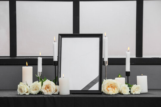 Empty black frame with a mourning ribbon, white candles, and roses on a dark table, funeral and remembrance