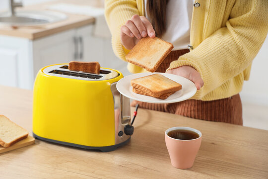Person making breakfast with a yellow toaster and serving toasted bread on a white plate with a cup of coffee, the person holds a slice of golden brown toast above a white plate with more toast