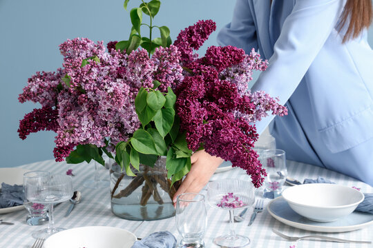 Woman arranging a fresh bouquet of purple and pink lilac flowers in a glass vase on a dining table