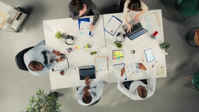 Office staff use laptops and computers from an overhead angle in a corporate workspace, using digital reports and charts to analyze information for office decisions.