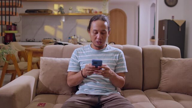 A man sits on a living room sofa using his smartphone, typing a message during a relaxed moment at home, illustrating everyday communication and modern digital lifestyle.