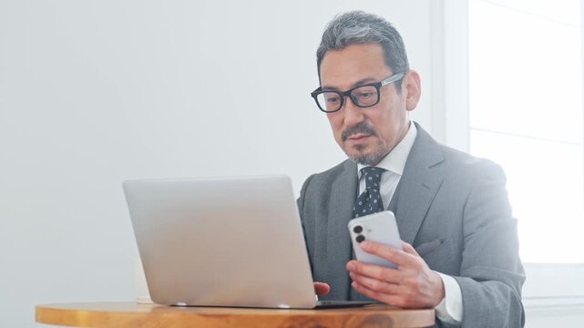スマホとノートPCを見るミドル男性。DX、カフェ、アプリ、ニュース、動画、リラックス、休憩、投資家