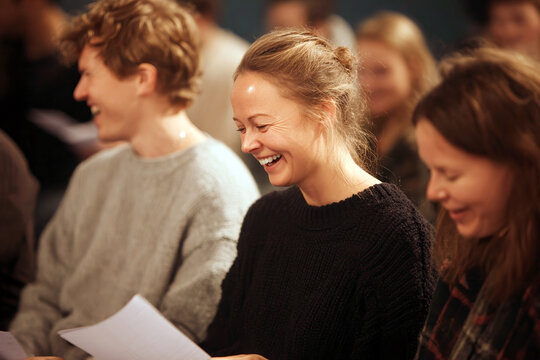 Group story swap participants sharing handwritten story sheets and laughing in a warm workshop setting for community event poster and promotion