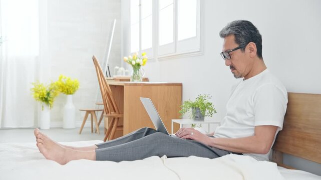 ベッドでノートPCを使うシニア男性。パソコン、仕事、DX、リラックス、ワーケーション、テレワーク