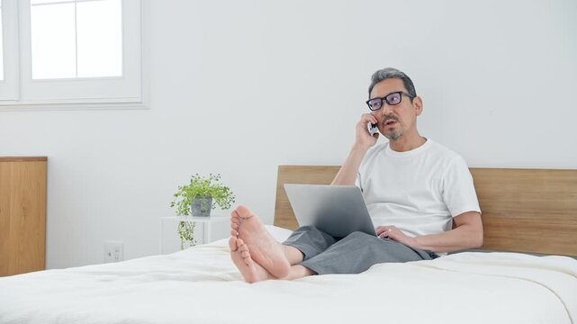 ベッドでノートPCを使うシニア男性。パソコン、仕事、DX、スマホ、通話、ワーケーション、テレワーク