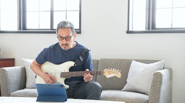 タブレットを見ながらエレキギターを弾く男性。音楽、楽器、演奏、練習、趣味、動画、バンド、音楽家、作曲