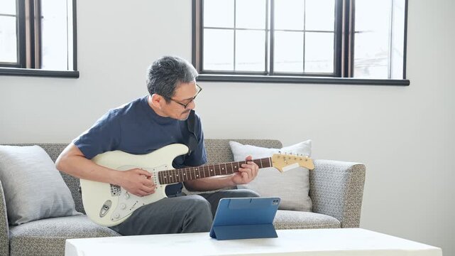 タブレットを見ながらエレキギターを弾く男性。音楽、楽器、演奏、練習、趣味、動画、バンド、音楽家、作曲
