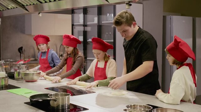 Young Caucasian male chef instructing group of Caucasian children rolling dough during culinary class. Video ideal for marketing children cooking workshops and educational programs