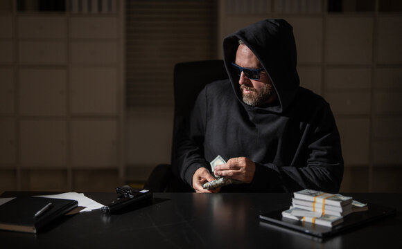 Robber counts stolen money indoors. Robber wears sunglasses and hoodie. Dangerous man appears as robber suspect. Handgun lies beside robber cash.