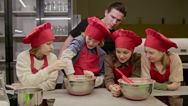 Caucasian teenagers wearing chef hats mixing ingredients in bowl during culinary class with male instructor. Perfect for promoting kids cooking workshops and educational programs