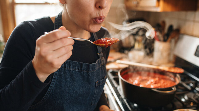 Woman tasting hot tomato sauce from a spoon in a home kitchen, blowing on the food to cool it down while wearing a denim apron