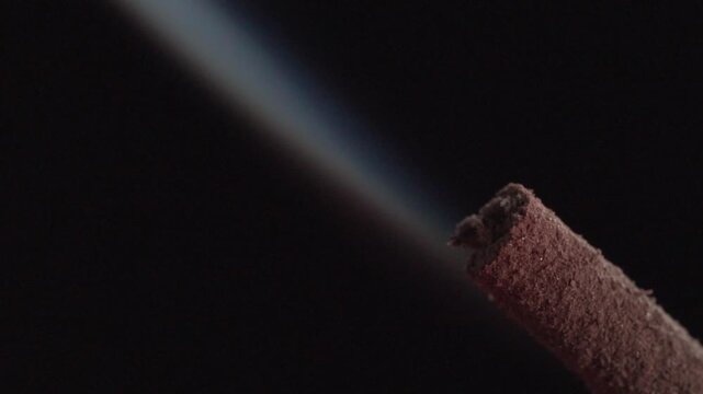 Macro view of smoldering incense stick releasing steady stream of smoke, highlighting burnt ash texture, combustion process, and atmospheric detail against dark background
