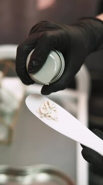 Vertical video. Cosmetic preparing. Skincare product. Professional beauty master hands dispensing white cream for woman treatment using spatula.