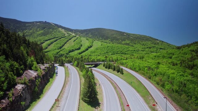 Amazing drone shot over Franconia Notch Parkway curves beneath Cannon Mountain as light traffic moves through lush green forest on a clear summer day, New Hampshire, United States