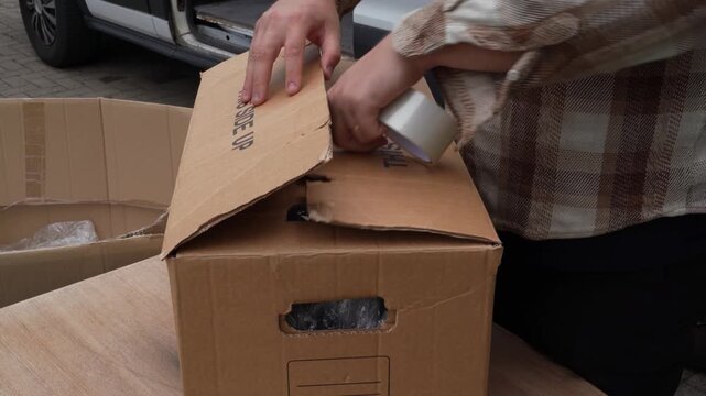 Open carton stuffed with loose old plastic wrap is closed and taped by a plaid-sleeved person as part of eco reuse shipping routine.