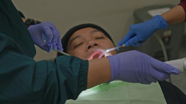 Dentist using UV curing light on male patient's teeth while assistant uses suction pipe