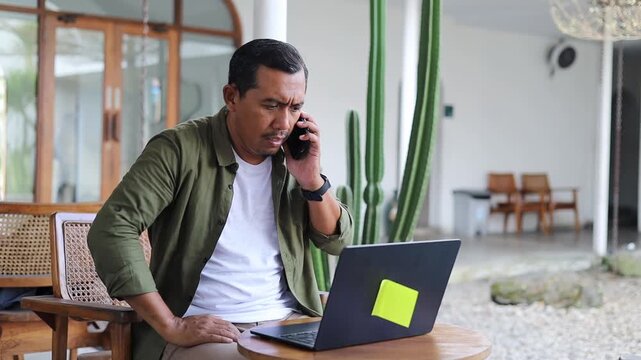 An adult man sits on a chair in a cafe, working on a laptop, angrily arguing with someone on a phone call.