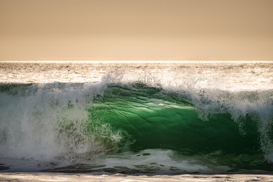 Emerald green ocean wave cresting with white foam under a golden sky