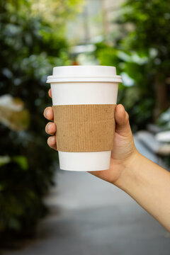 Hand holding a white takeaway coffee cup with a green nature background