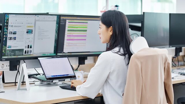 A young professional woman in a white shirt working at a computer workstation in a bright modern open-plan office with city views.