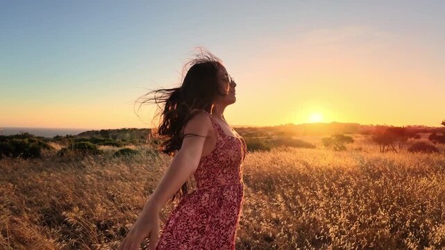 A young woman stands with arms outstretched, feeling happy and free in a beautiful golden field as the sun sets. Her long hair blows gently in the wind, embracing the peaceful nature.