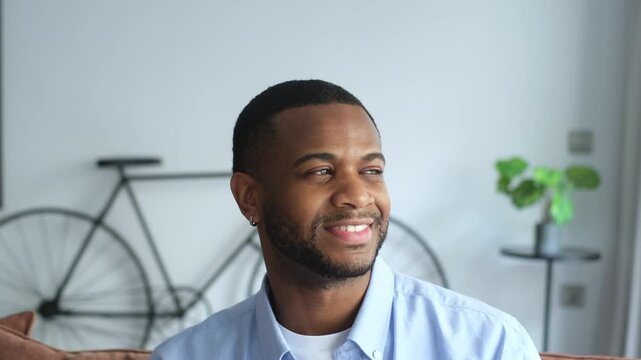 A cheerful African American man sits comfortably in a modern indoor setting, looking contentedly into the distance before turning to smile warmly at the camera. He exudes positivity and confidence.