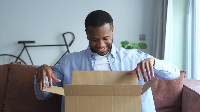 A happy young African American man joyfully opens a cardboard package at home, excited about his online order. He smiles as he discovers its contents.