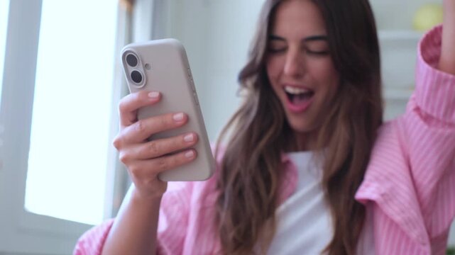 Surprised and overjoyed young woman using her smartphone, receiving amazing good news, celebrating a success or a victory, and expressing genuine happiness with an excited yes gesture at home