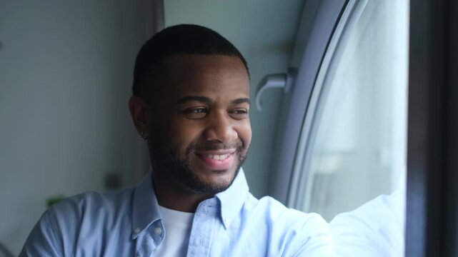 Hopeful young african american man standing by the window, looking outside with a contemplative smile, feeling optimistic and inspired while enjoying the view from his modern home