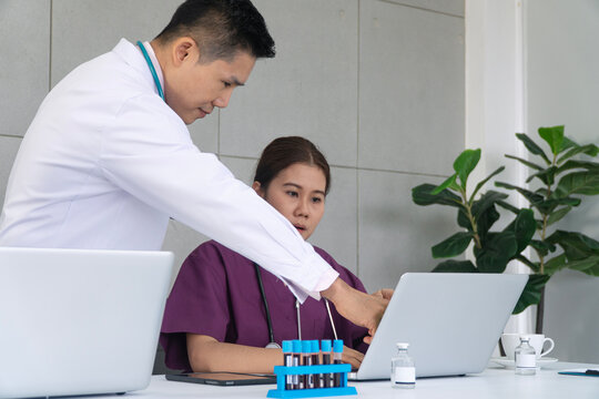 Asian male doctor and female nurse discussing patient blood test results on laptop in clinic. Medical team working together in hospital laboratory consulting digital health records.