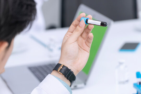 Close up over shoulder view of a male doctor holding a blood sample test tube in hospital laboratory. Scientist analyzing patient medical data on laptop with green screen for mockup replacement.