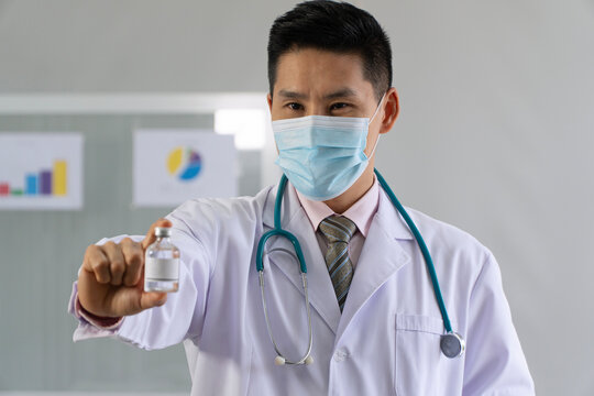 Asian male doctor in medical face mask holding a glass vial of vaccine or medicine with a blank label. Professional physician presenting a new treatment, cure, or immunization in clinic.