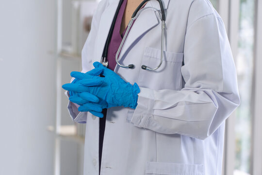 Close up of unrecognizable medical professional in white lab coat and blue sterile gloves with clasped hands. Confident doctor or nurse standing in bright hospital clinic ready for patient care.
