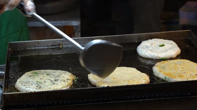 Close-up of a street vendor frying traditional roti flatbread with green onions on a hot griddle surface at a night market, slow motion. Thai street food concept