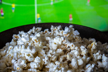 Obraz premium A delicious bowl of salty popcorn in focus with a green football field showing an active sports game blurred on a television screen at home in Brazil.