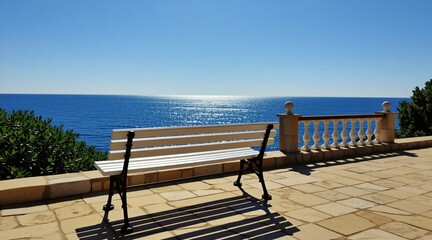 Obraz premium Empty white wooden bench stands on stone terrace overlooking Mediterranean sea. Sunny day brings clear blue sky, calm water horizon. Relaxing place for vacation travel leisure. Scenic outdoor
