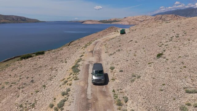 Silver camper van driving a dusty gravel road along the coastline of Pag Island, Croatia. Aerial