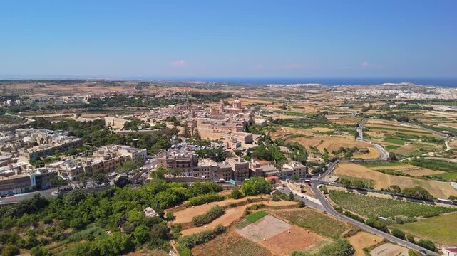 Mdina, Malta fortified medieval city with St Paul's Cathedral, bastions and historic rooftops amid countryside fields under clear summer skies, captured in an aerial wide shot.