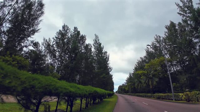 POV shot driving down the dual carriageway between Victoria and the airport on Mahe, Seychelles. Lush tropical trees line the island's only highway under an overcast sky.