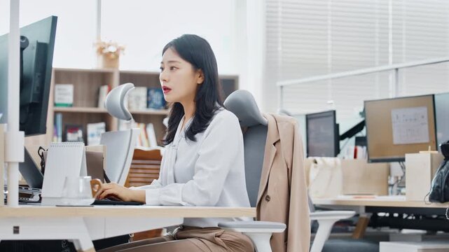 A young professional woman sitting at a workstation in a bright modern office with large windows and city views working on a computer.