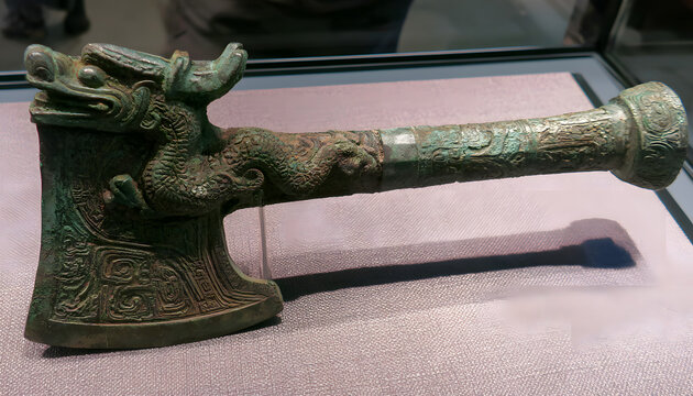 An ancient bronze axe from the Chinese Bronze Age, adorned with a dragon motif. The axe head and handle feature intricate carvings. Represents an artifact from the Shang Dynasty era. Displaying cultur