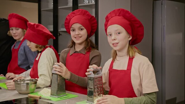 Slowmo portrait of Caucasian female teenagers wearing red chef hats and aprons, holding graters in kitchen. Footage is perfect for promoting culinary schools or children cooking classes