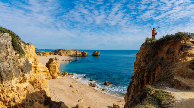 happy woman with raised arms standing on cliff looking at beach in algarve portugal