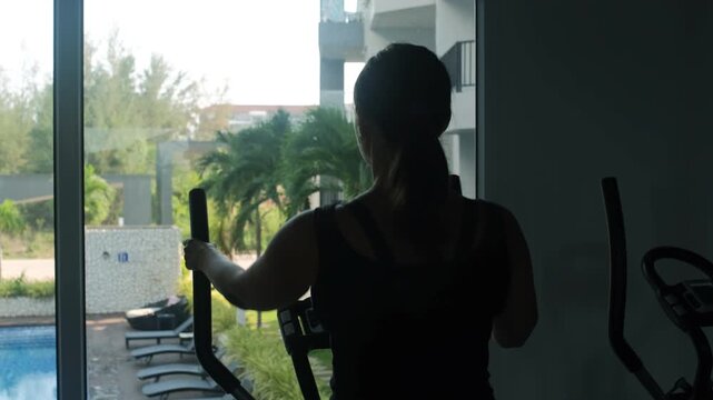 Dramatic dark silhouette of a young woman using an elliptical trainer in a gym with strong backlight from large windows with pool and palms outside, slow motion. Fitness concept