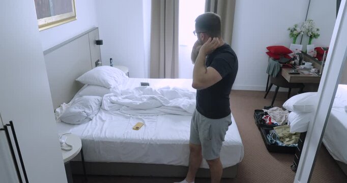 Man scratching his body repeatedly while standing next to a hotel bed. Travel concept of itchy skin, possible bed bug bites, accommodation hygiene concerns, and guest discomfort.