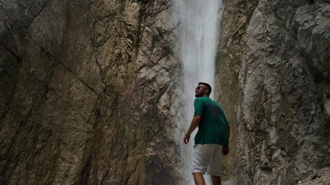 A young man stands close to a waterfall inside a narrow rocky gorge. Water drops behind him between steep stone walls.