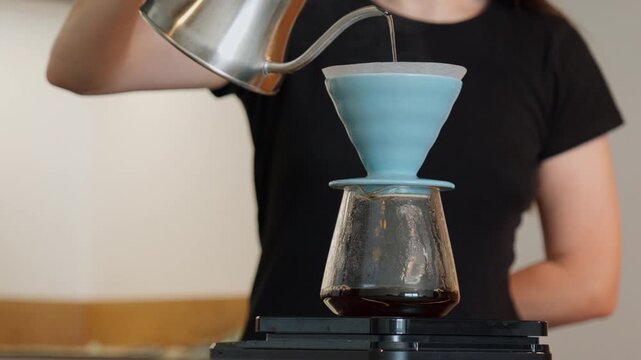 A woman pours hot water from a gooseneck kettle into a blue dripper with a paper filter. Fresh brew drips into a glass server on a kitchen counter.